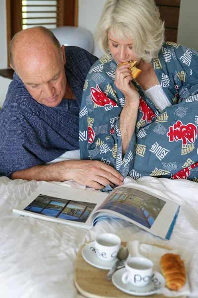 Older man and woman in bathrobe sitting on a bed taking their breakfast while le