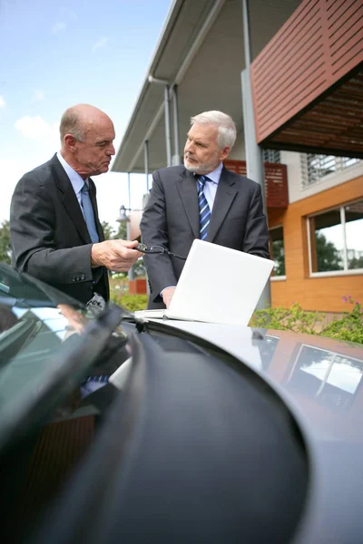 Older men in suits in front of a laptop sitting on the hood of a car