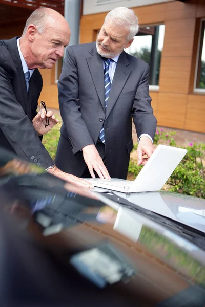 Older men in suits in front of a laptop sitting on the hood of a car