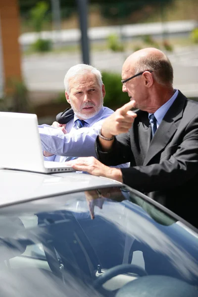 Older men in suits in front of a laptop computer placed on the roof of a car