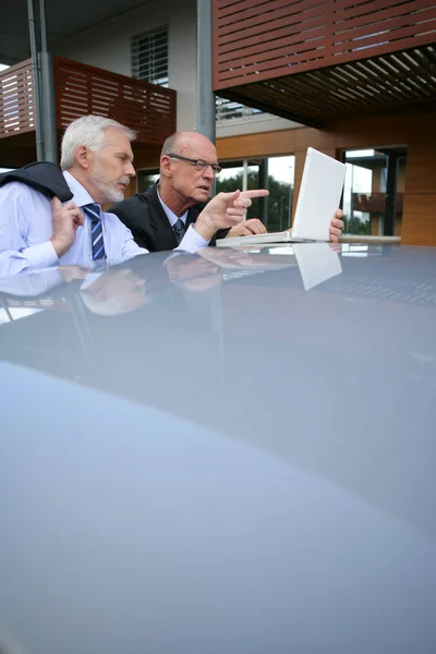 Older men in suits in front of a laptop computer placed on the roof of a car