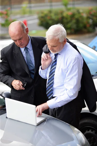 Older men in suits in front of a laptop sitting on the hood of a car