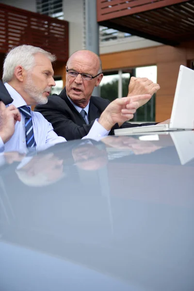Older men in suits in front of a laptop computer placed on the roof of a car