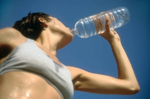 一个穿着运动服的女人的低视角 在蓝天的映衬下喝着水瓶里的水 横向射击 — 图库照片