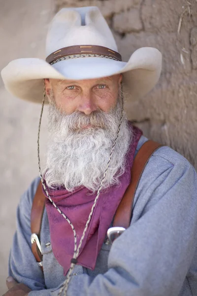 Portrait of a cowboy wearing a tall hat and sporting a long white beard. He is dressed in a heavy work shirt and kerchief. Vertical shot.