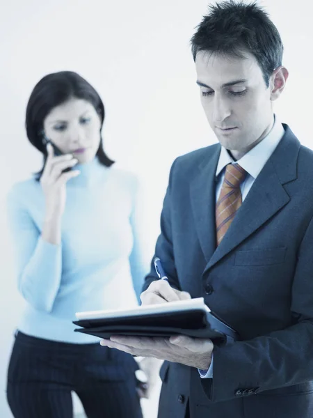 Businessman writing on a personal organizer with a businesswoman standing behind him holding a mobile phone