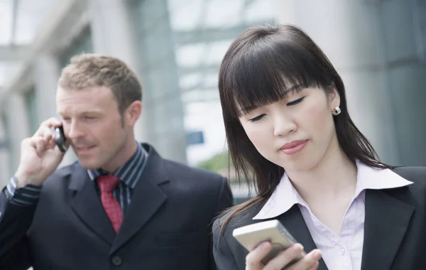 Close-up of a businesswoman using a personal data assistant with a businessman talking on a mobile phone behind her
