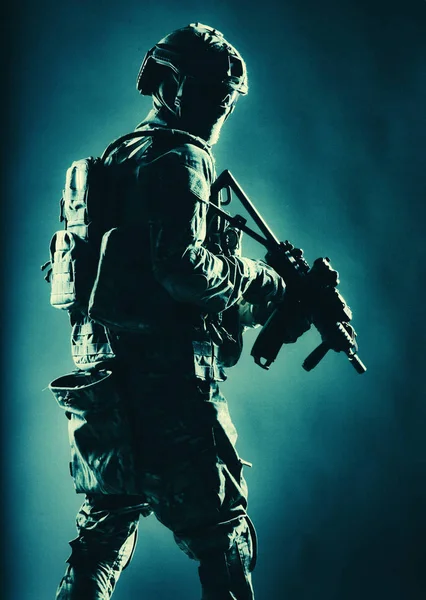 Army soldier in helmet, battle uniform with tactical ammunition, hiding face behind mask and glasses, looking back over shoulder, sneaking in darkness with service rifle in hand, low key, studio shoot