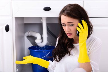 Sad Young Woman Calling Plumber In Front Of Water Leaking From Sink Pipe clipart
