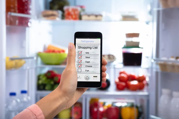 Close-up Of A Woman\'s Hand Showing Shopping List On Mobile Phone In The Refrigerator