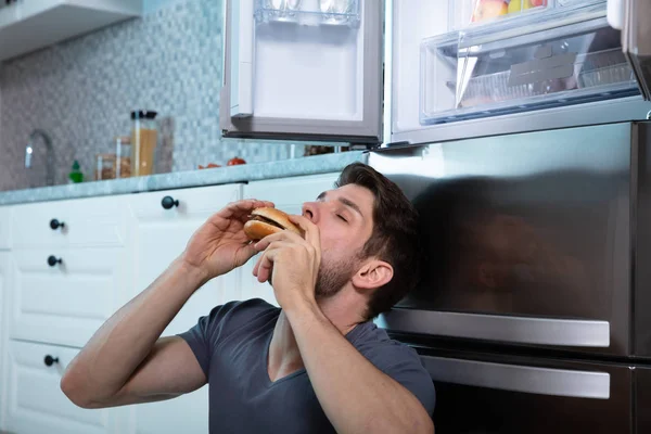 Közelkép Egy Férfiról Aki Hamburgert Eszik Ahogy Hűtőszekrényre Támaszkodik — Stock Fotó