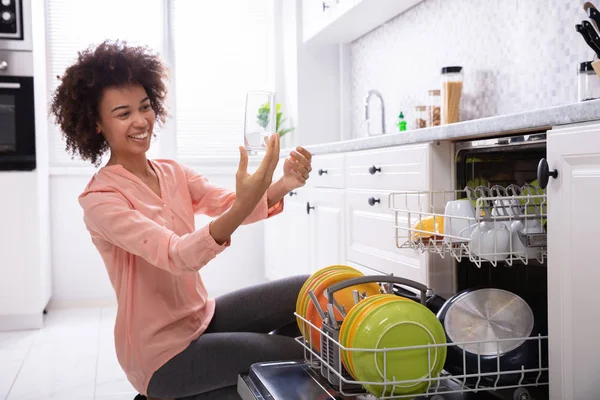 幸せな若い女チェック洗浄食器洗い機に近い透明なコップ ロイヤリティフリーのストック画像