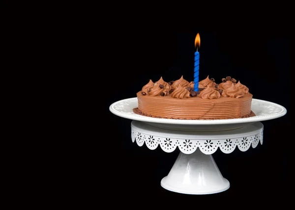 Home made chocolate cake with chocolate fudge on top on an off white porcelain plate with blue candle burning brightly on a white pedestal and black background with copy space