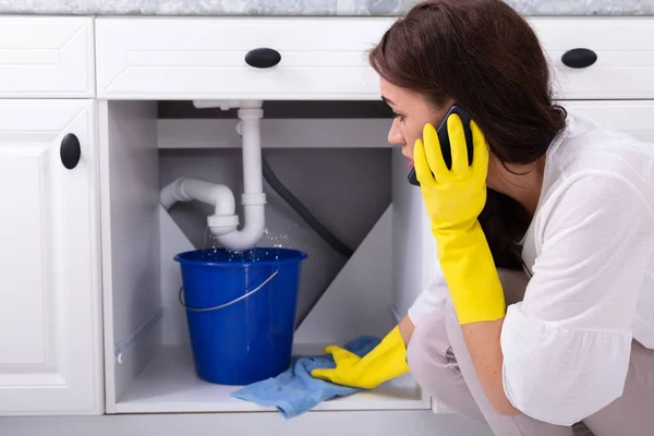 Sad Young Woman Calling Plumber In Front Of Water Leaking From Sink Pipe