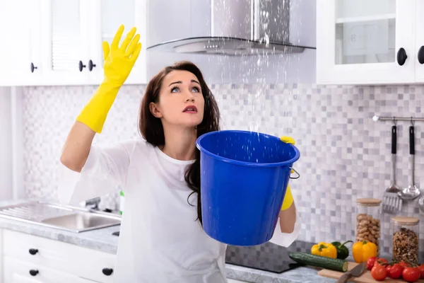 不快乐的年轻女子收集漏水从天花板在蓝色桶 — 图库照片