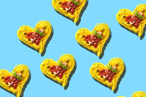 Decorative hearts of pasta of cooked yellow spaghetti with tomatoes, basil and parmesan cheese toping, viewed from above in full frame on light blue background