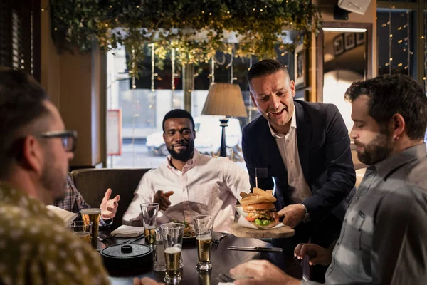 Mid adult waiter placing a meal down on the table where a small group of mid adult men are sitting and relaxing.