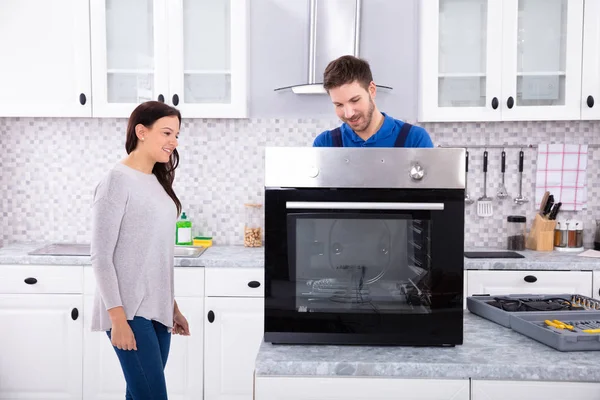 Smiling Repairman Repairing Oven On Kitchen Worktop In Front Of Woman