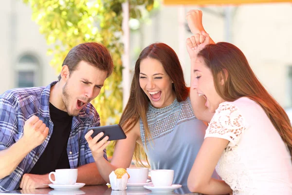 Three excited friends celebrating good news watching smart phone media content in a coffee shop