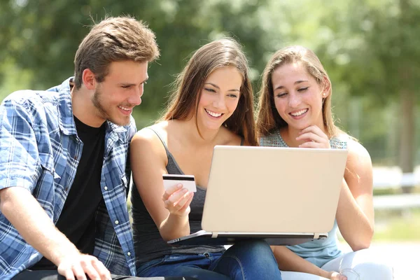 三个快乐的朋友坐在公园的长椅上 用信用卡和笔记本电脑在网上交钱 — 图库照片
