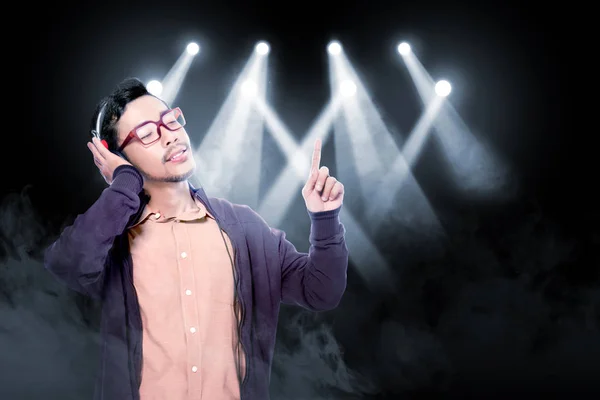 愉快的亚洲人在眼镜穿着棕色夹克听音乐用耳机在烟和音乐会现场照明背景 — 图库照片