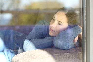 Sad woman looking through a window lying on a couch in a house interior clipart