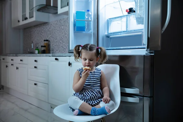 Cute Little Girl Eating Cupcake While Sitting Front Open Refrigerator — стоковое фото