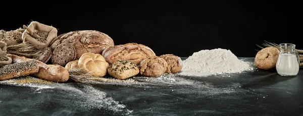 Panorama display of gourmet breads with dough, milk and flour ingredients over a floured kitchen table with copy space
