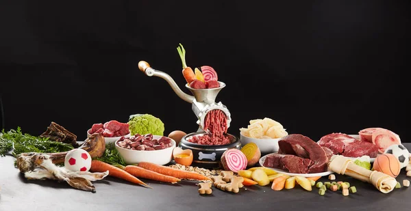 Ingredients for preparing BARF food for dogs. Meat with vegetables mincing in grinder, bones, fresh beef and greens arranged on kitchen table against black background