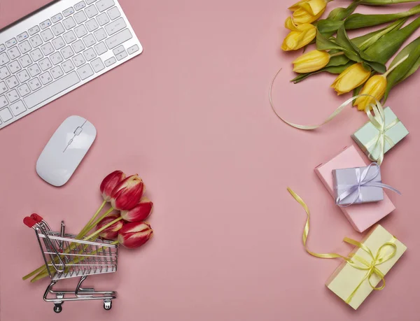 Shopping trolley, gift boxes, tulips flowers, computer keyboard and mouse on pink background. Top view with copy space. Valentine's day, Mother's day, Women's day, wedding. Seasonal sale