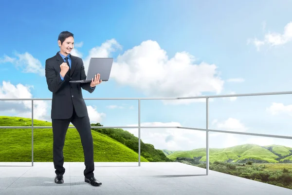 Successful asian businessman standing with laptop on the terrace with green landscapes view and blue sky background