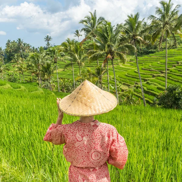 Relaxed fashionable caucasian woman wearing red asian style kimono and traditional asian paddy hat walking amoung beautiful green rice fields and terraces on Bali island.