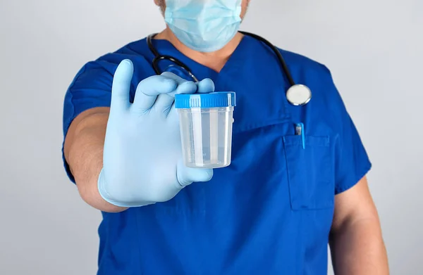 医生身穿蓝色制服 乳胶手套拿着一个空塑料容器 取尿样 — 图库照片