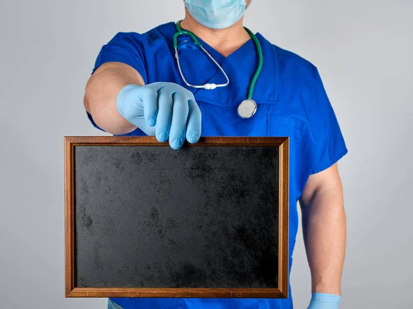 医生在蓝色制服和无菌乳胶手套拿着一个木框架与空白黑色的文字背景 灰色的背景 — 图库照片