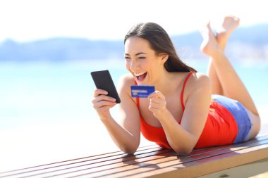 Excited girl finding ecommerce offers buying online with phone and credit card on the beach clipart