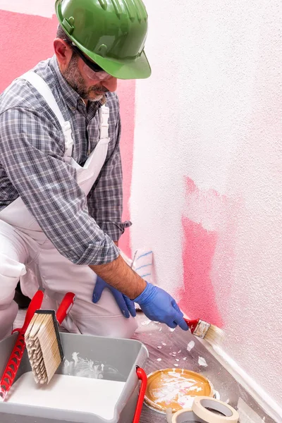 白い全体に白人の家の画家の労働者は ヘルメットとゴーグルは小さなブラシを使用して白い塗料でピンクの壁を塗装します 建設業 作業安全 — ストック写真
