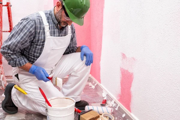 白いオーバーオールの白人の家の画家の労働者は ヘルメットとゴーグルで 彼はピンクの壁をペイントするために白い塗料を準備します 建設業 作業安全 — ストック写真