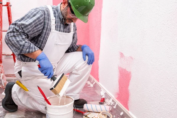 白いオーバーオールの白人の家の画家の労働者は ヘルメットとゴーグルで 彼はピンクの壁をペイントするために白い塗料を準備します 建設業 作業安全 — ストック写真