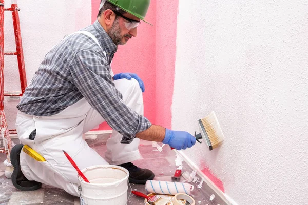 白い全体に白人の家の画家の労働者は ヘルメットやゴーグルは ブラシを使用して白い塗料でピンクの壁を塗装します 建設業 作業安全 — ストック写真