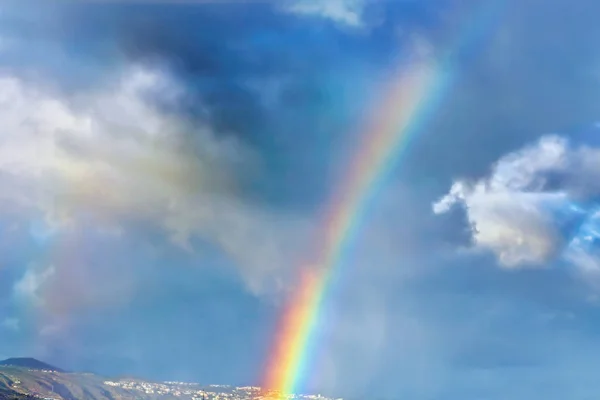 ほぼ垂直方向の虹の間にいくつかの白い雲が青空に遠くにジャンプします 写真の下にはテネリフェ島のカナリア島の村が見えます — ストック写真