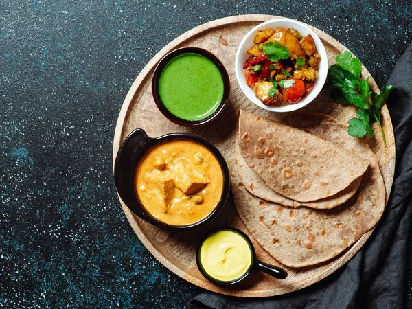 Indian cuisine dishes: vegetable curry, shahi paneer, chapati, chutney. Indian food on wooden tray over dark background. Assortment indian meal with copy space for text. Top view or flat lay.