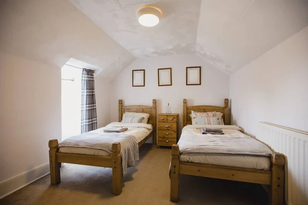 Interior shot of a empty bedroom. There are two single wooden beds against a wall with a set of drawers between them.