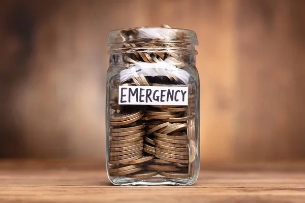 Coins In Jar With Emergency Label Over Wooden Desk
