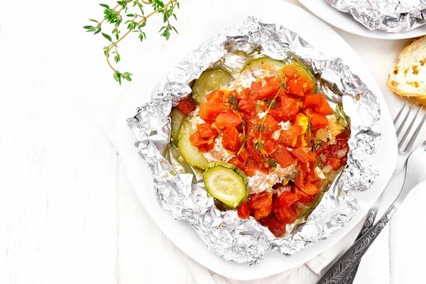 Pink salmon with zucchini, tomatoes, onions, garlic and thyme, baked in foil on a plate, napkin, fork and bread on a wooden board background from above