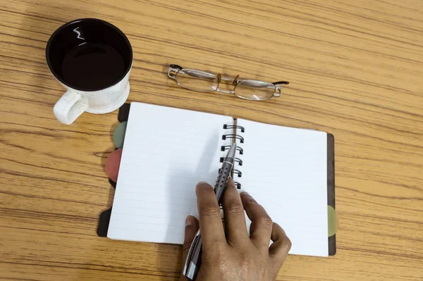 ビジネスマンは彼の日記に書いている 木製のテーブルの上にコーヒーと眼鏡のカップの横に空白のページでノートブックを開きます ビジネスはまだテーブルの上のオフィスの事と生命概念 コピースペース付きのトップビュー — ストック写真