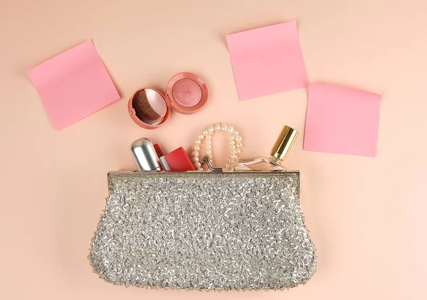 open silver clutch and cosmetics fell out of the middle, red lipstick,  peach background, top view