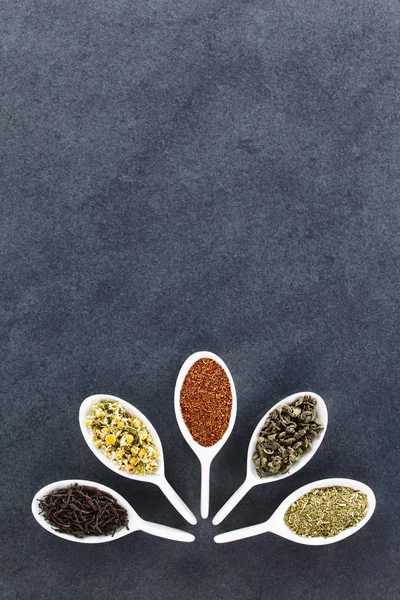 A variety of loose dried tea leaves on spoons (black, chamomile, rooibos, green and mate tea), photographed overhead on slate with copy space on the top