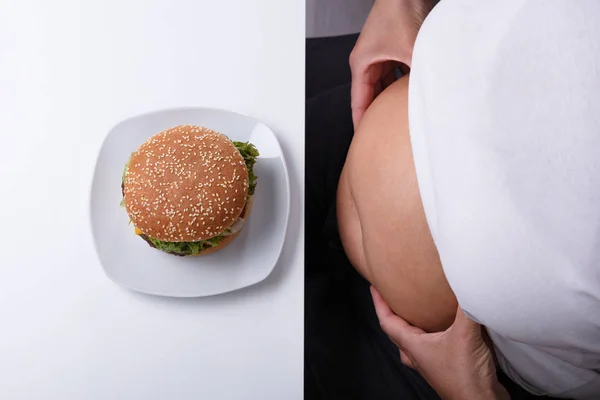 An Overhead View Of Woman Pinching Her Fatty Stomach With Hamburger On Plate