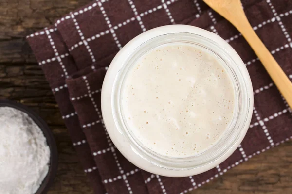 Fresh homemade bubbly sourdough starter, a fermented mixture of water and flour to use as leaven for bread baking, in glass jar with flour and wooden spoon on the side, photographed overhead (Selective Focus, Focus on the top of the starter)