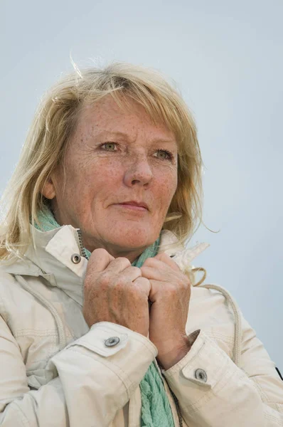 upper body portrait of a blond mature woman in bright jacket in front of cloudy sky dreaming to the side looking up with hands on collar